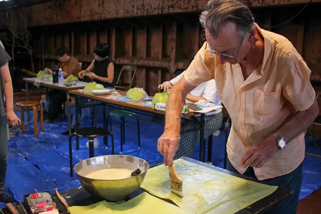 クロード・ロシュさんが、仮面型に貼り付ける紙に、小麦粉糊を塗っている。